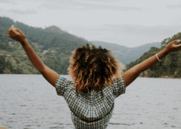 Une jeune femme noire pose de dos les bras en l'air. Elle est sur le bord d'un lac et a une posture inspirée et confiante.