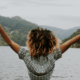 Une jeune femme noire pose de dos les bras en l'air. Elle est sur le bord d'un lac et a une posture inspirée et confiante.
